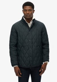 Mørk quiltet jakke med høj krave, med trykknapper og kontrasterende kant, båret over en grå sweater.