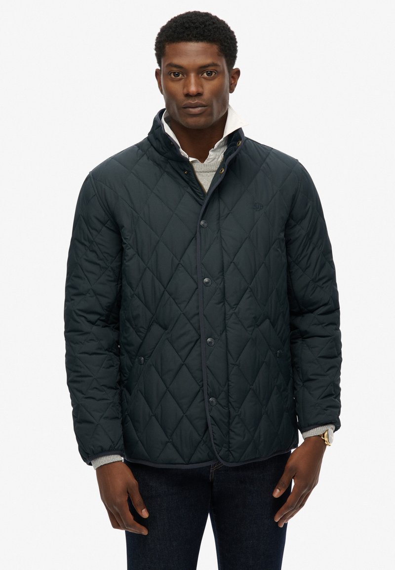 Mørk quiltet jakke med høj krave, med trykknapper og kontrasterende kant, båret over en grå sweater.