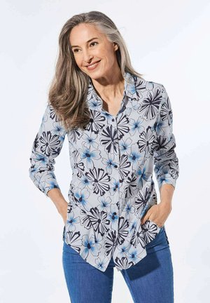 Camicia a maniche lunghe con bottone a fiori, caratterizzata da uno sfondo grigio chiaro, grandi fiori blu e neri e orlo curvo. Indossata con jeans blu.