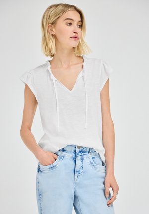 Lyshåret kvinde iført en hvid kortærmet bluse med flæsekant og lyseblå højtaljede jeans, den ene hånd i lommen.