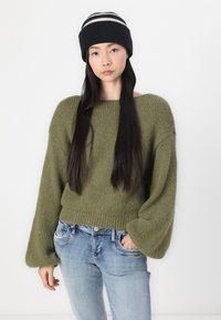 Olivgrüner Strickpullover mit weiten Ärmeln, kombiniert mit hellblauen Jeans. Schwarze Strickmütze mit weißen Streifen.