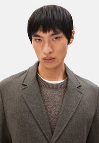 Grau melierter Blazer mit Fischgrätmuster und Reverskragen, über einem strukturierten braunen Strickpullover getragen. Nahaufnahme, neutraler Hintergrund.