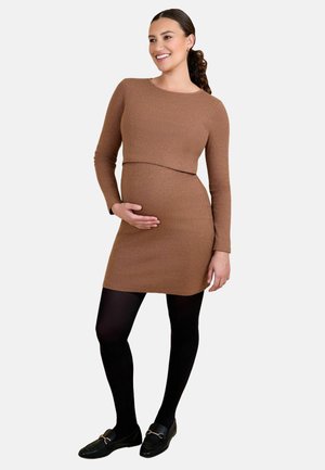 Schwangere Frau in einem braunen, figurbetonten Kleid, schwarzen Strumpfhosen und schwarzen Loafers, lächelnd und hält mit einer Hand ihren Bauch.