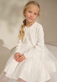 Robe blanche avec jupe en tulle et cardigan brodé, avec des manches longues et des bords festonnés. Tissu lisse, texture superposée et détails délicats.