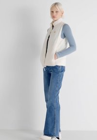 White insulated vest over a light blue long-sleeve top, paired with blue flared jeans and white sneakers. Minimalistic design, no visible patterns.