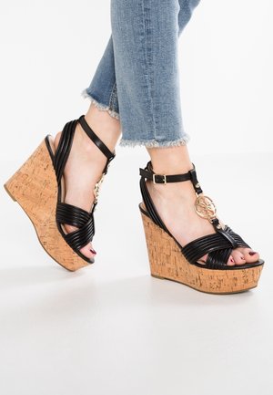 Sandalias de cuña negras con base de corcho, diseño de correas entrelazadas, acento de herrajes dorados y puntera descubierta. Combinadas con jeans de denim azul deshilachados.