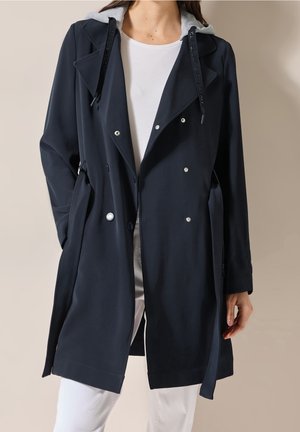 Femme portant un trench-coat noir croisé avec des boutons argentés et une capuche grise, associé à un pantalon blanc et une chemise blanche.