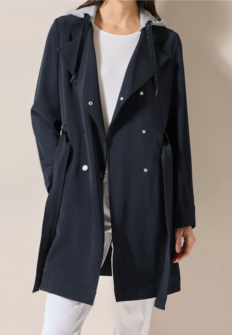 Femme portant un trench-coat noir croisé avec des boutons argentés et une capuche grise, associé à un pantalon blanc et une chemise blanche.