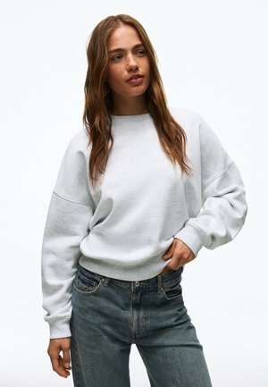 Vrouw met lang bruin haar die een lichtgrijze oversized trui en blauwe spijkerbroek draagt, poseert met één hand op haar heup.