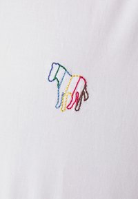 Forme animale abstraite colorée brodée sur fond de tissu blanc, avec des contours en fils bleu, vert, jaune, rouge et marron.
