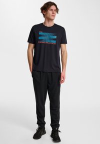 Zwarte T-shirt met korte mouwen met blauw en oranje "PERFORMANCE" grafisch ontwerp, gecombineerd met zwarte sweats en zwarte sportschoenen.