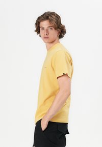 Gul t-skjorte i bomull med korte ermer, rund hals og lite brodert logo på brystet. Kombinert med svarte shorts med glatt tekstur.