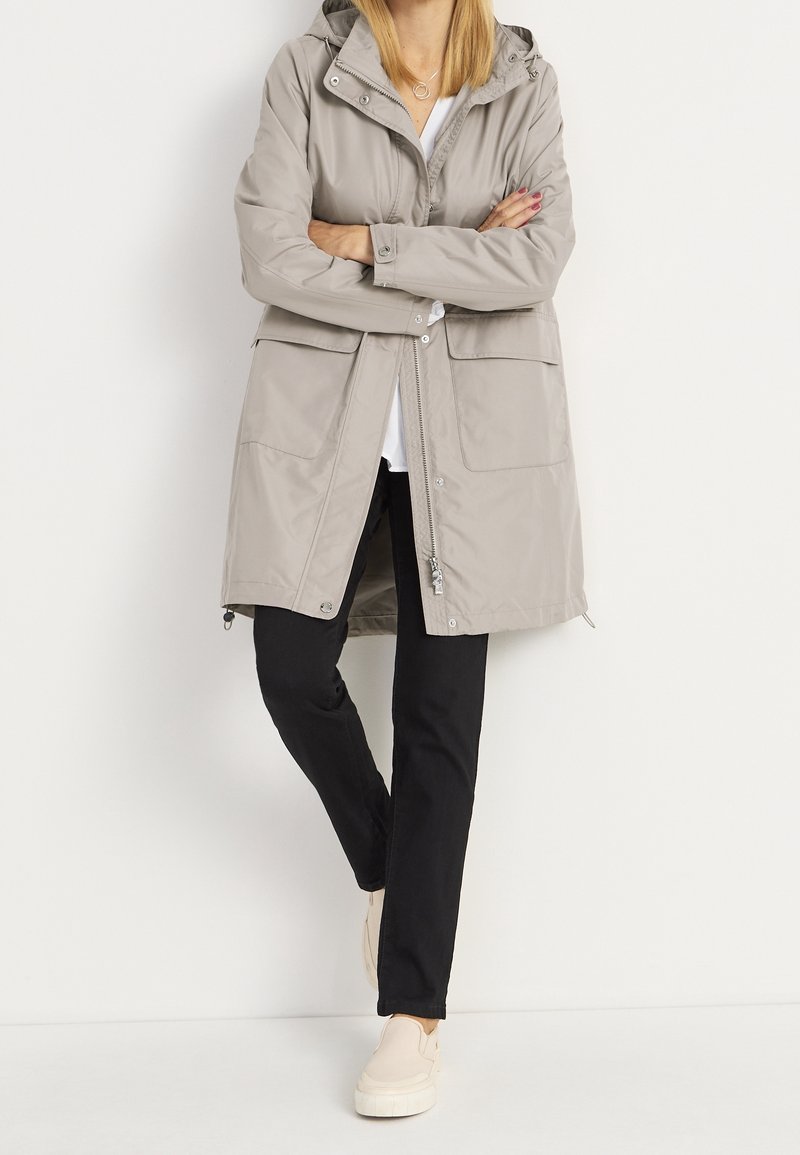 Vaalea harmaa vesitiivis parka hupulla, jossa on kaksi etutaskua, vetoketjusuljenta ja säädettävä helma, yhdistettynä mustiin housuihin ja vaaleisiin kenkiin.