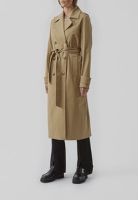Beige trenchcoat lavet af et glat stof, med dobbeltradet design, bælte i taljen og store revers. Fremhævet med knapper.