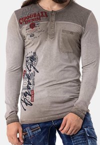 Camiseta henley de manga larga en gris claro con paneles en los hombros de gris más oscuro, con un bolsillo frontal y gráficos de texto superpuestos en rojo y negro.