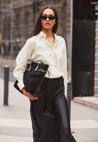 Chemise boutonnière couleur crème, jupe en satin noir et sac en suède noir avec des accents en cuir. Des lunettes de soleil noires surdimensionnées complètent le look.