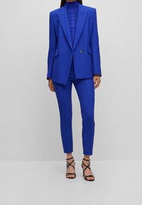 Jasnoniebieska, dopasowana marynarka z szerokimi klapami, zapinana na jeden guzik, zestawiona z dopasowanymi, wąskimi spodniami i czarnymi, paskowymi szpilkami.