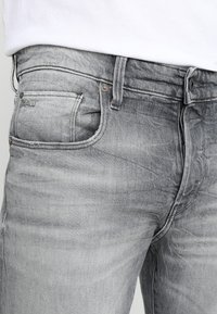 Los jeans de mezclilla gris presentan un ligero desvanecimiento, un diseño de cinco bolsillos y acentos de hardware metálico. La tela tiene un acabado suave y texturizado.