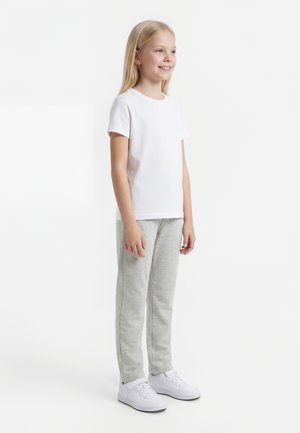 Girl with long blonde hair smiling, wearing a plain white t-shirt, light gray sweatpants, and white sneakers, standing against white background.