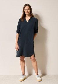 Marineblaues, knielanges Kleid mit V-Ausschnitt und langen Ärmeln. Hergestellt aus strukturiertem Stoff mit seitlichen Schlitzen, kombiniert mit weißen Sneakers.
