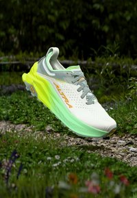 Λευκό και γκρι αθλητικό παπούτσι με νέον πράσινη και κίτρινη σόλα, με την ένδειξη «ALTRA», να αιωρείται πάνω από ένα μονοπάτι από χαλίκια και γρασίδι.