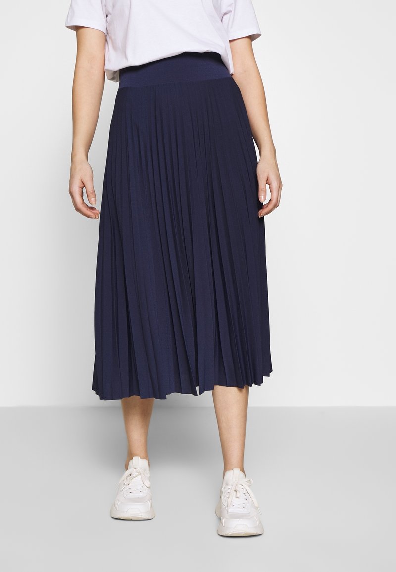 Personne portant une jupe midi plissée bleu marine, un t-shirt blanc et des baskets blanches, debout devant un fond blanc uni.