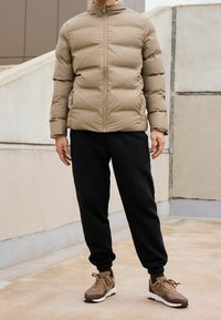 Beżowa kurtka puchowa z wysokim kołnierzem, czarne spodnie dresowe i brązowe sneakersy, charakteryzujące się teksturowanym wzorem i zapięciem na zamek.