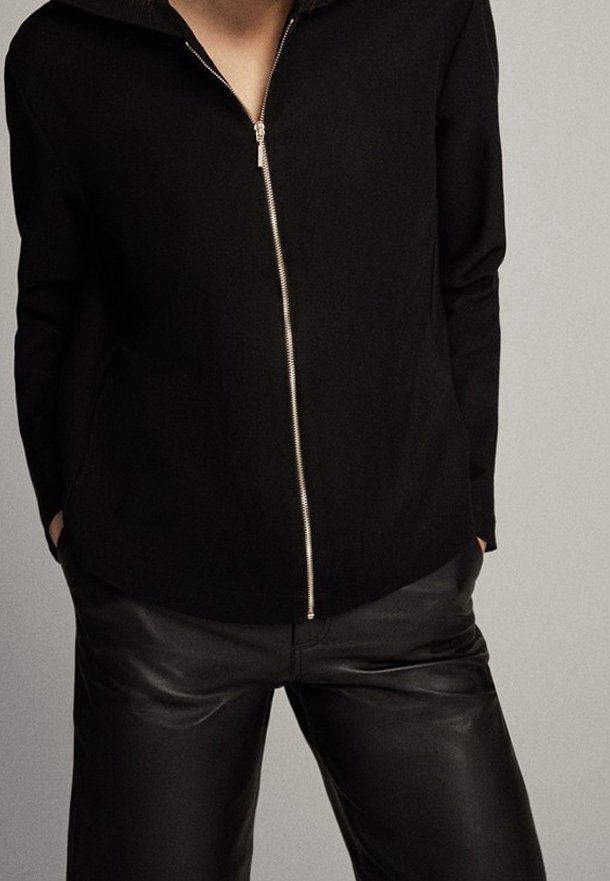Zwarte ritssluitingjack met hoge kraag, voorzien van een zilveren rits en lange mouwen, gecombineerd met nauwsluitende zwarte leren broek.