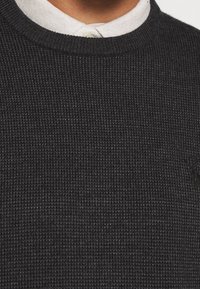 Suéter negro texturizado con cuello redondo. Presenta un patrón de tejido sutil y está superpuesto sobre una camisa blanca con cuello.