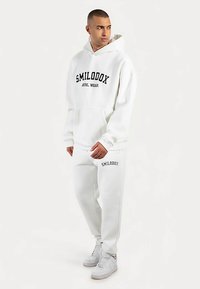 Witte hoodie en bijpassende joggingbroek, beide met "SMILODOX" in het zwart bedrukt. Zachte, comfortabele stof met een ontspannen pasvorm.