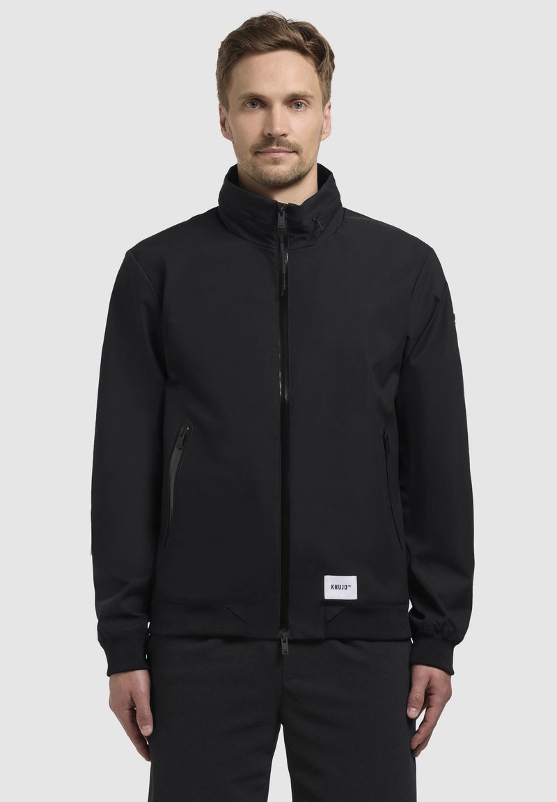 Chaqueta negra con cremallera, cuello alto, bolsillos laterales y puños acanalados. Confeccionada en tejido suave con una etiqueta de logo en el dobladillo.