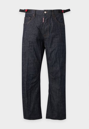 Homme portant un t-shirt marron, un jean foncé avec une fermeture éclair latérale, des bottes noires, une ceinture avec une boucle colorée et une casquette marron et blanche, debout devant un fond uni.