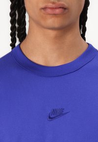 Nike purpurs krekls ar apaļu kakla izgriezumu un īsām piedurknēm. Tam ir izšūts logs atbilstošā krāsā kreisajā krūtīs. Gluda tekstūra.