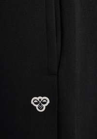 Schwarzer Stoff mit einer glatten Textur, der ein gesticktes Logo in Weiß und Schwarz zeigt. Das Design beinhaltet ein dezentes Nahtdetail an der Seite.