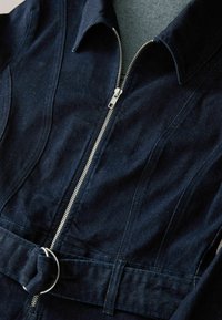 Dunkelblauer Denim-Overall mit einem Reißverschluss vorne, spitzem Kragen und einem Stoffgürtel mit einem Metallring. Glatte Textur mit strukturierten Nähten.