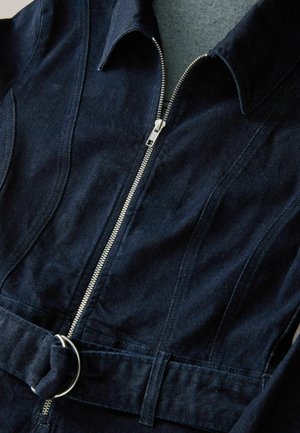 Marineblauwe denim jumpsuit met een ritssluiting aan de voorkant, puntige kraag en een stoffen riem met een metalen ring. Soepele textuur met gestructureerde naden.