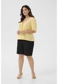 Cardigan en tricot jaune avec un décolleté en V et des manches courtes, fermeture à boutons, associé à un short noir et des chaussures à talons jaunes.