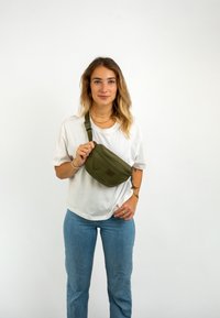 Olíva zöld deréktáska sima felülettel, cipzáras záródással és állítható pánttal. Fehér ing felett, kék farmernadrággal viselve.