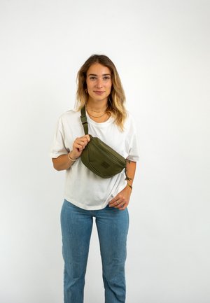 Olivengrüne Gürteltasche mit glatter Oberfläche, Reißverschluss und verstellbarem Tragegurt. Trägt man über einem weißen Hemd mit blauen Jeans.
