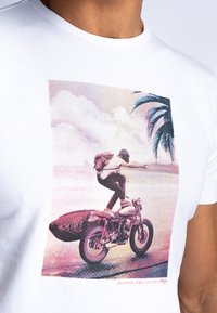 Bela bomba majica iz bombaža z grafičnim tiskom osebe na motociklu s surfboards, palmami in pisano ozadjem zahoda.