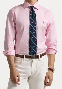 Camicetta rosa chiaro con maniche arrotolate, abbinata a una cravatta blu navy con strisce verdi diagonali e piccoli dettagli dorati, indossata con jeans bianchi.