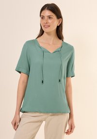Top mint green a maniche corte con scollatura a V, dettaglio con cordoncino e trama sottile, abbinato a pantaloni beige chiaro.