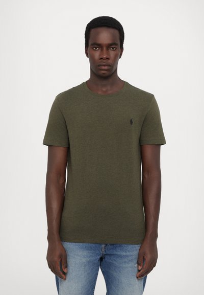 T-shirt en coton vert olive avec un col rond et des manches courtes, arborant un petit logo noir sur la poitrine gauche. Porté avec un jean bleu clair.