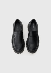 Černé kožené mokasíny s tlustými, vroubkovanými gumovými podrážkami. Obsahují dvojité kožené pásky přes nárt a jemné detaily prošívání.