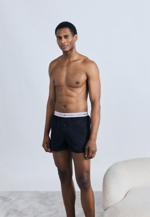 Μαύρα βαμβακερά μποξεράκια με γκρι ζωνάκι στη μέση που δείχνει το λογότυπο "TOMMY HILFIGER"· το μοντέλο είναι γυμνό ανώμαλο και στέκεται μπροστά σε ανοιχτόχρωμο φόντο.