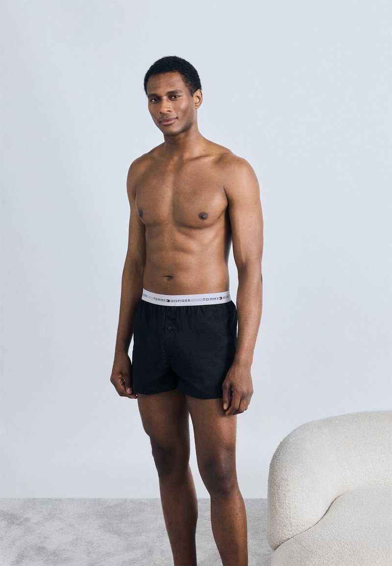 Μαύρα βαμβακερά μποξεράκια με γκρι ζωνάκι στη μέση που δείχνει το λογότυπο "TOMMY HILFIGER"· το μοντέλο είναι γυμνό ανώμαλο και στέκεται μπροστά σε ανοιχτόχρωμο φόντο.