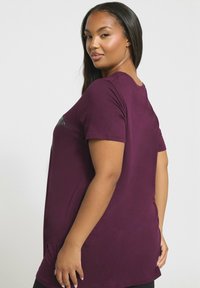 Nainen, jolla on pitkät tummat hiukset, pukeutuneena purppuraan lyhythihaiseen paitaan, seisoo hieman sivuttain yksinkertaista taustaa vasten.