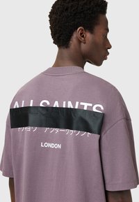 Camiseta de manga corta gris-púrpura con un panel de satén negro y texto en relieve en la parte posterior, así como una marca blanca encima. Ajuste casual.