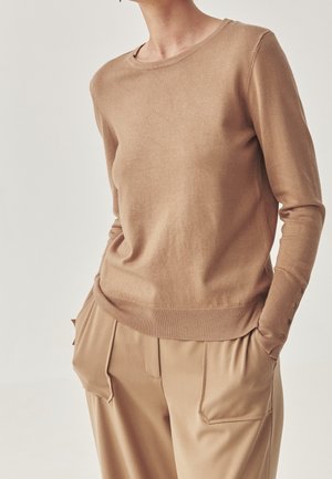 Personne portant un pull beige à manches longues avec des détails de boutons sur les poignets et un pantalon beige assorti avec de grandes poches avant.
