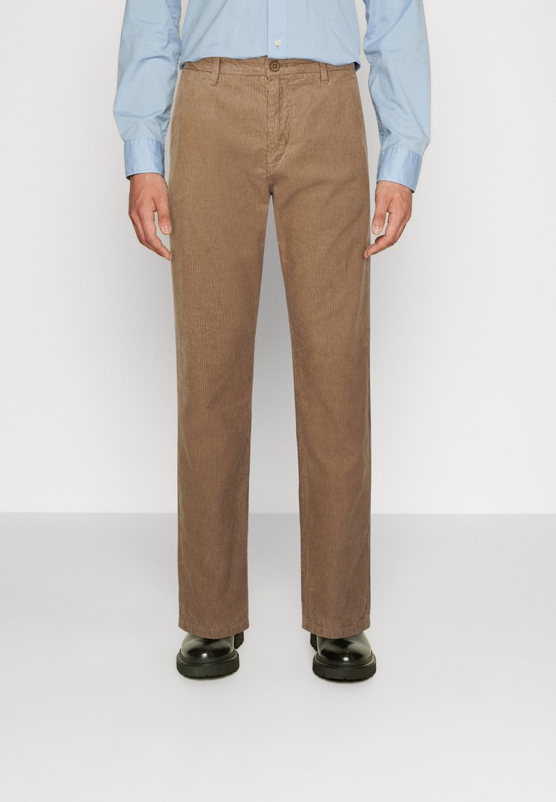 Homme portant une chemise bleu clair, un pantalon en velours côtelé marron et des chaussures noires, debout devant un fond uni.